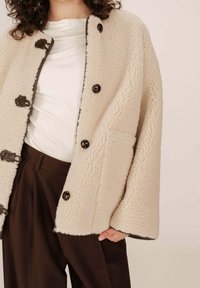 Cappotto in pile beige con una superficie testurizzata, bottoni neri e rifiniture marroni. Indossato sopra una maglietta bianca e pantaloni marroni.