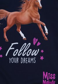 Illustrazione di un cavallo marrone in movimento su un tessuto blu navy, con "Segui i Tuoi Sogni" in bianco e accenti di glitter rosa, accanto a "Miss Melody."