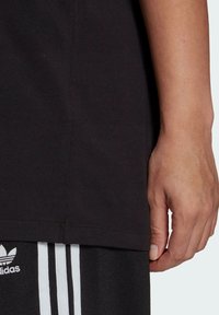 Černé bavlněné tričko s krátkým rukávem, s rovný lem a viditelným bílým logem blízko spodního okraje. Nosič má na sobě černé kalhoty s bílými pruhy.