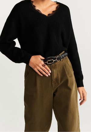 Femme portant un pull noir à col en V avec bordure en dentelle, un pantalon taille haute olive, une ceinture noire et une bague à la main gauche.