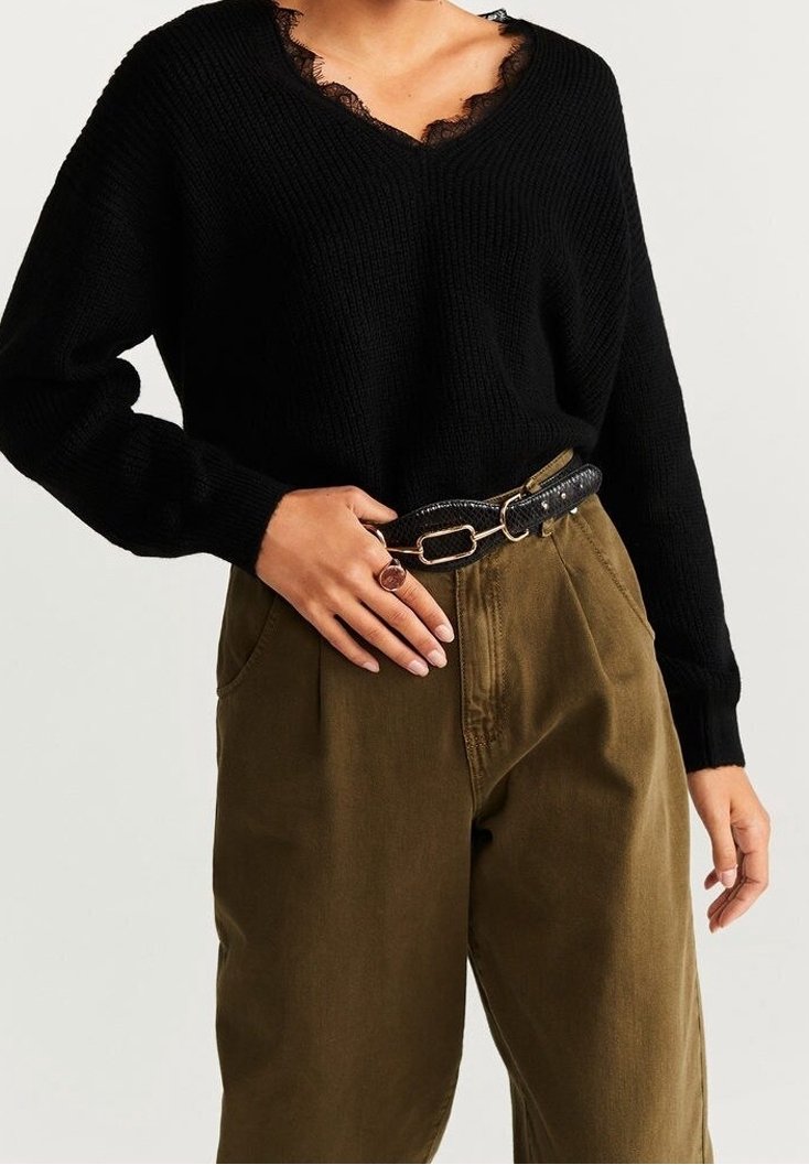Femme portant un pull noir à col en V avec bordure en dentelle, un pantalon taille haute olive, une ceinture noire et une bague à la main gauche.