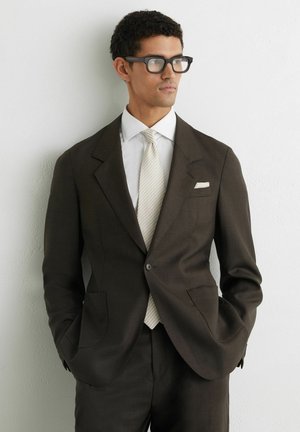 Mand iført mørkebrun jakkesæt, hvid skjorte og lyst stribet slips, stående med hænderne i lommerne mod en hvid væg, kiggende mod højre.