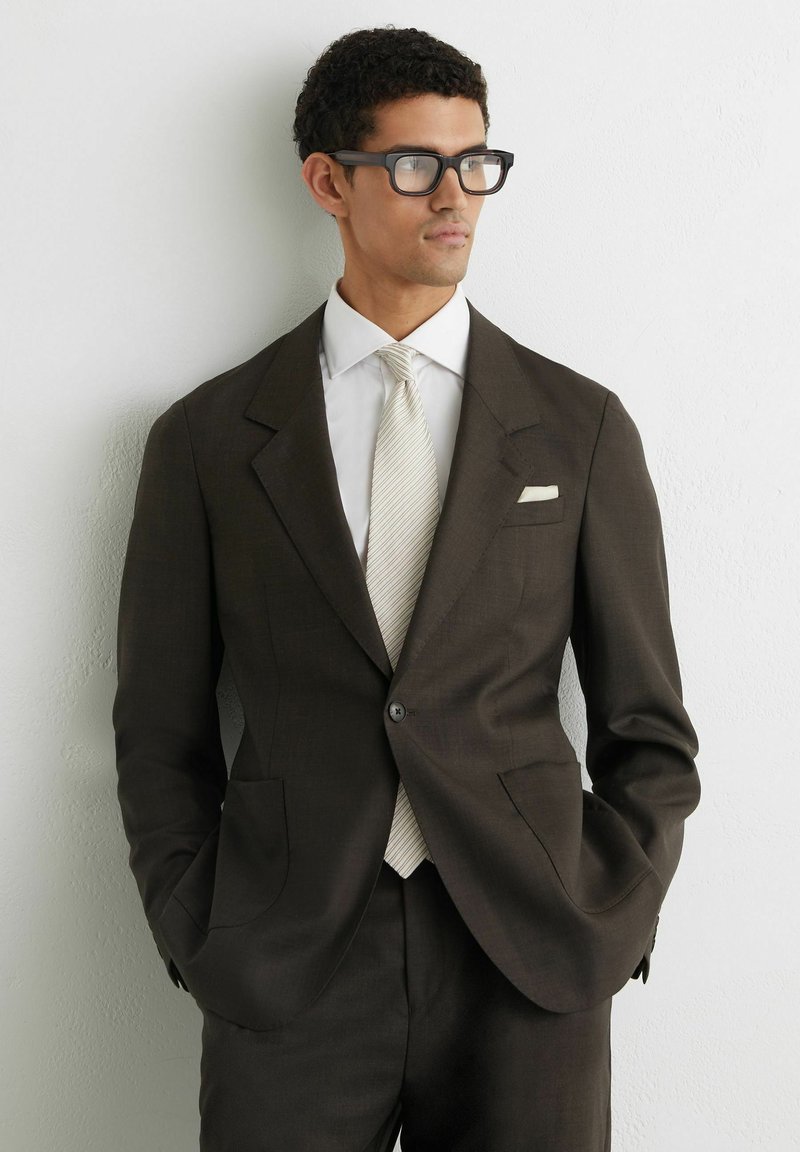 Mand iført mørkebrun jakkesæt, hvid skjorte og lyst stribet slips, stående med hænderne i lommerne mod en hvid væg, kiggende mod højre.