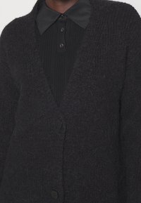 Svart stickad cardigan med texturerad yta, med V-ringning, lång ärm och tre framknappar. Bärs över en svart ribbad topp.