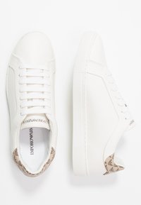 Vita läder sneakers med beige varumärkesflik på hälen, snörningsdesign och tjock gummisula visad från ovan och sidan.