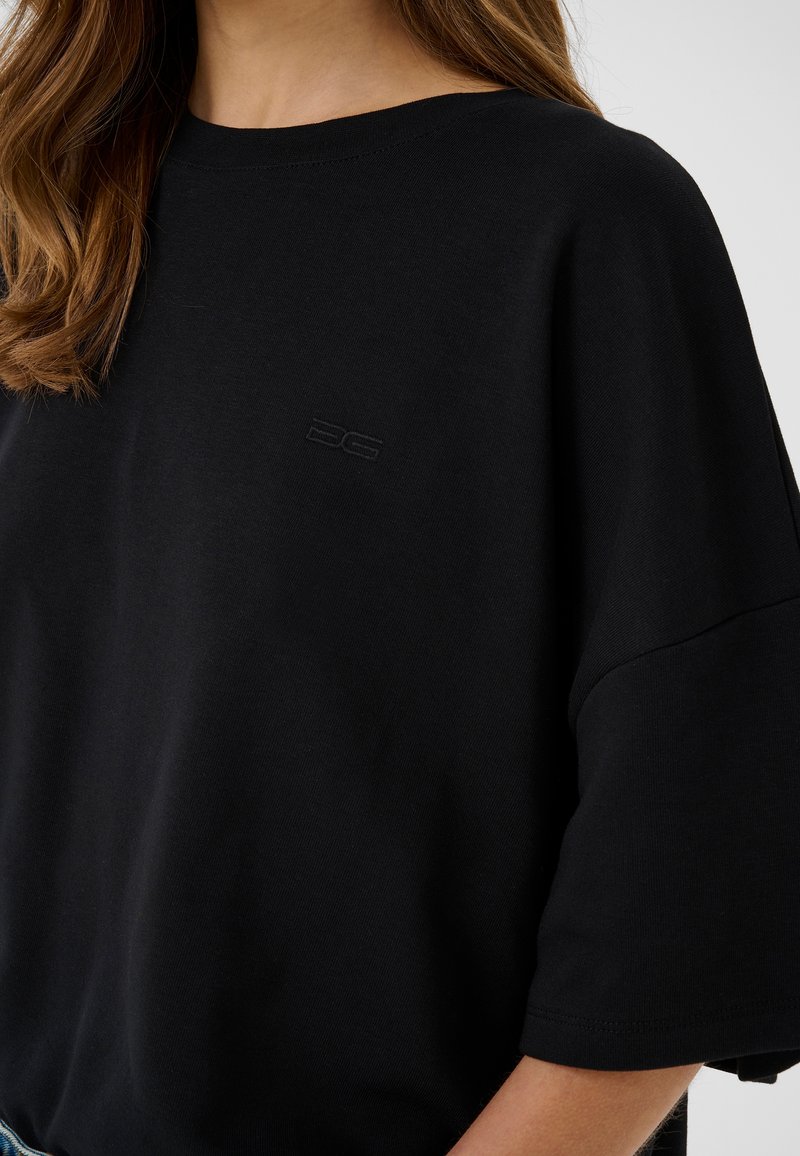 Femme portant une chemise noire ample à manches courtes avec un petit logo embossé sur la poitrine et des cheveux ondulés châtain clair.
