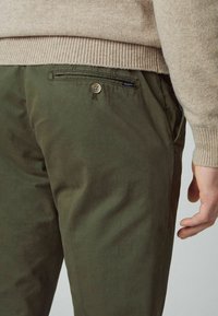 Gros plan sur une personne portant un pantalon vert olive avec une poche arrière boutonnée et un pull beige en maille, la main reposant le long du corps.