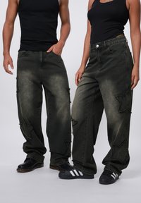 Baggy, dark green jeans with star-shaped cutouts and frayed edges. Worn with black shoes; textured fabric visible. Plain background.