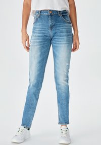 Augsti vidukļa, gaiši zilas džinsu bikses ar pieguļošu piegriezumu, kurās ir nolietotas vietas un klasiskā piecu kabatu dizains. Iekļautas baltas sporta kurpes.