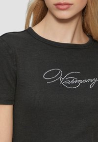 Schwarzes, geripptes T-Shirt mit Rundhalskragen und kurzen Ärmeln, das ein dekoratives silbernes Strassschriftlogo auf der Brust trägt.