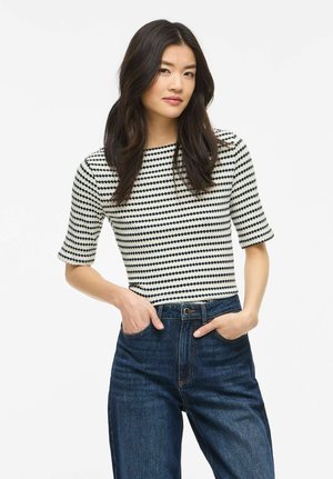 Giovane donna con lunghi capelli scuri indossa una maglia a pois bianca e nera e jeans blu a vita alta, con le mani nelle tasche, su uno sfondo bianco.