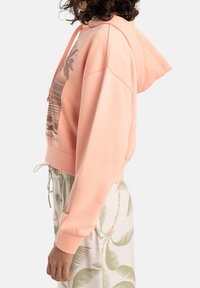 Sudadera corta de color rosa claro con capucha, que presenta un estampado gráfico en la parte delantera y un dobladillo con cordón, combinada con pantalones de color claro y con patrón.