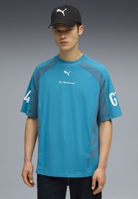 Turkosa t-shirt med kort ärm och grå detaljer, med en vit Puma-logotyp och BMW Motorsport-märken på bröstet och ärmarna.