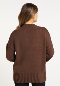 Barna kötött pulóver magas nyakkal, laza fazonnal, oldalsó hasítékokkal és bordázott aljjal. A textúrája lágy és tömör.