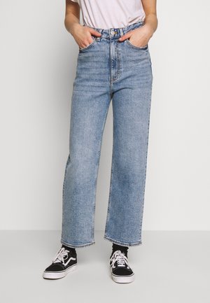 Henkilö, jolla on yllään vaaleansiniset korkeavyötäröiset leveälahkeiset farkut, valkoinen t-paita, joka on tungettu housuihin, kädet taskuissa, mustat sukat ja mustat lenkkarit valkoisilla nauhoilla.