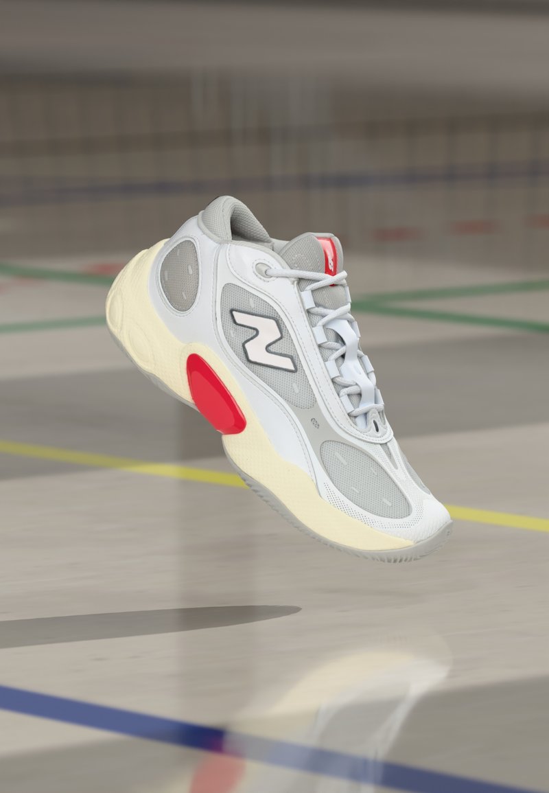 Λευκό και γκρι αθλητικό παπούτσι με κόκκινες λεπτομέρειες να αιωρείται πάνω από το δάπεδο εσωτερικής αθλητικής γηπέδου με ζωγραφισμένες γραμμές.