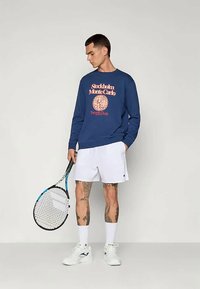 Jūras spēku kreklis ar "Stokholma Montekarlo" grafiku, apvienots ar baltām sporta šortiem un turot tenisa raketi. Gluda auduma, brīva piegriezuma.