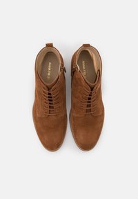 Botas de tobillo de ante marrón con punta redondeada, cordones en la parte delantera, detalles de cremallera lateral y una superficie texturizada. Nombre de la marca visible en el interior.