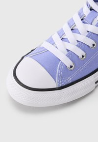 Ανοιχτό μωβ υφασμάτινο αθλητικό παπούτσι με λευκά κορδόνια, στρογγυλεμένο λευκό κάλυμμα μύτης και μαύρο περιθώριο στη σόλα από καουτσούκ. Διαθέτει μεταλλικά οπές για κορδόνια και υφή στην επιφάνεια.