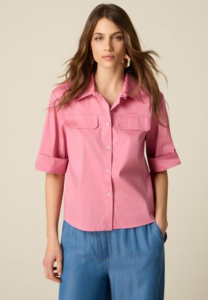 Kvinde med løst brunt hår iført en pink skjorte med knapper og opkærmede ærmer og blå bukser, stående mod en ensfarvet beige baggrund.