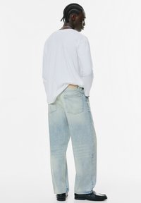 Jeans decolorate într-o nuanță de albastru deschis, cu o croială confortabilă, având buzunare la spate și o textură estompată, purtate împreună cu o cămașă albă cu mânecă lungă.