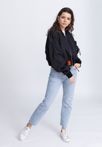 Black bomber jacket with oversized fit, zipper closure, and orange accent. Light blue straight-leg jeans and white sneakers.