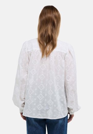 Blouse blanche en broderie anglaise avec motifs en losange, manches longues bouffantes et boutons au poignet. Portée avec un jean bleu.