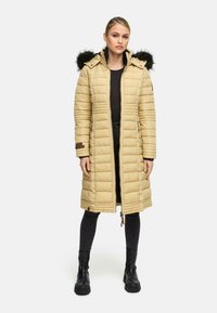 Polstret lang frakke i lys beige med hætte i pels, lodret quilting og lynlås. Matcher med sorte jeans og kraftige sorte støvler.