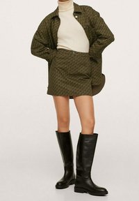 Chemise vert olive avec un motif géométrique, assortie à une mini-jupe coordonnée et des bottes hautes en cuir noir. Design simple et ajusté.