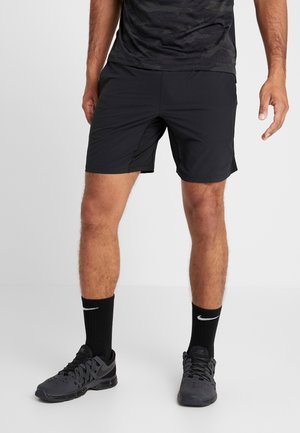 Mężczyzna ubrany w czarne sportowe spodenki, czarne skarpetki Nike oraz czarne buty sportowe Nike, stojący na tle białego, gładkiego tła.