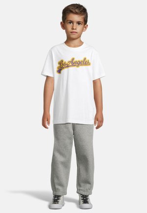 T-shirt en coton blanc avec imprimé "Los Angeles" en violet et jaune, associée à un pantalon de survêtement gris et des baskets grises avec des lacets noirs.