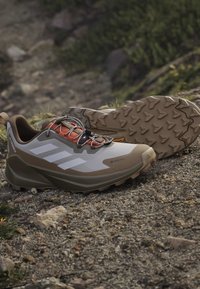 Ζευγάρι καφέ και μπεζ παπούτσια πεζοπορίας Adidas Terrex Gore-Tex σε βραχώδες έδαφος, που δείχνουν τη σόλα και την πλευρά με τις τρεις λευκές ρίγες.