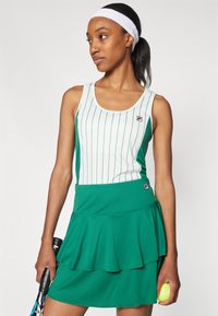 Grön tennisklänning med vita vertikala ränder, lagerkjol och Fila-logotyp. Modellen håller en tennisracket och boll, och bär ett vitt hårband.