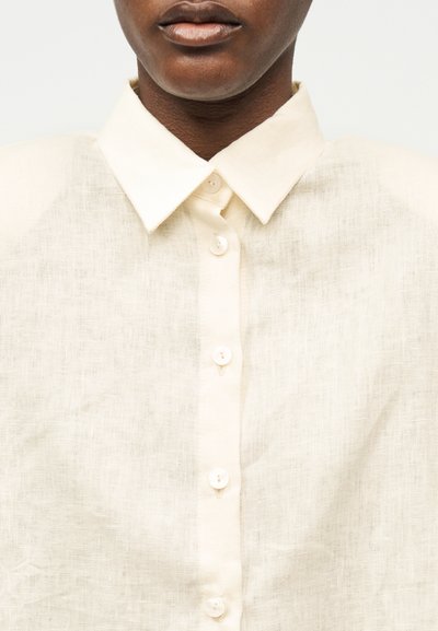 Κοντινό πλάνο ενός ατόμου που φοράει μια λινή πουκάμισα σε εκρού χρώμα με μυτερό γιακά και ορατά λευκά κουμπιά κατά μήκος του μπροστινού μέρους.