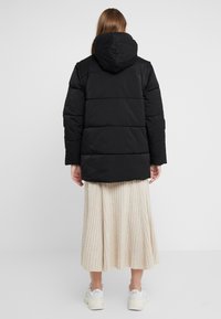 Chaqueta puffer negra con capucha, con secciones acolchadas y un corte holgado, llevada sobre una falda plisada de color beige claro y zapatillas blancas.