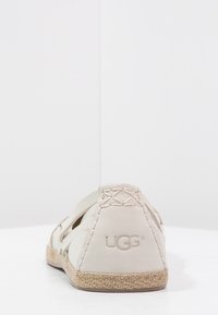 Zapato plano beige UGG con suela trenzada y detalle de costura en el talón, visto desde la parte posterior sobre una superficie blanca contra un fondo blanco.