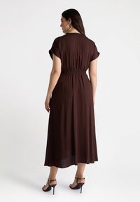 Braunes, leichtes Midikleid mit kurzen Ärmeln und gesmocktem Taillendetail. Getragen mit schwarzen High Heels und Creolen.