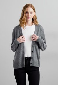 Grå cardigan laget av myk stoff, med V-hals, ribbede mansjetter og seks svarte knapper foran, sammen med en hvit skjorte.