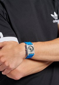 Montre avec un bracelet en silicone bleu et un cadran rectangulaire argenté, des aiguilles des heures et des minutes argentées, et un motif circulaire noir au centre.