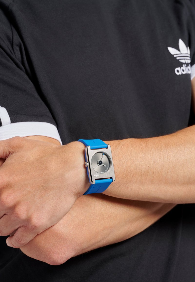 Montre avec un bracelet en silicone bleu et un cadran rectangulaire argenté, des aiguilles des heures et des minutes argentées, et un motif circulaire noir au centre.