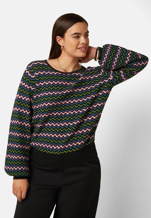 Joven con cabello castaño liso lleva un jersey de manga larga con patrón de zigzag multicolor y pantalones negros, posando con la mano detrás de la cabeza.