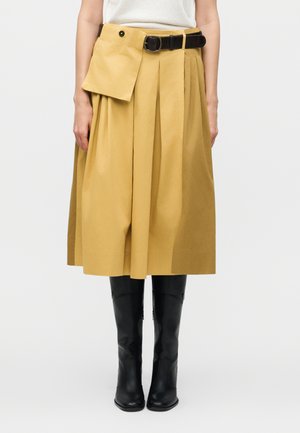 Žena nosí plisovanú midi sukňu horčicovej farby s pripevnenou klopovou vreckou a čiernym opaskom, skombinovanú s čiernymi čižmami po kolená.