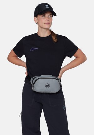 Vrouw met een zwarte Mammut-pet, een zwart Mammut-T-shirt en marineblauwe broek met een grijze Mammut-heuptas, staand met haar handen in haar zij.