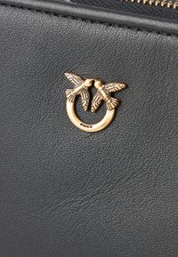 Bolsa de couro texturizado preto com um fecho de metal dourado em forma de duas aves. Nome da marca "PINKO" visível na ferragem.