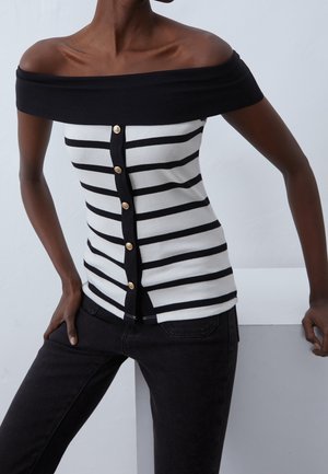 Persoon draagt een zwart-wit gestreepte off-shoulder blouse met knopen, gecombineerd met een zwarte broek, staand met handen in de zij.