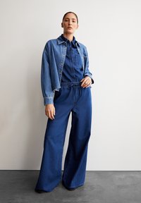 Denimoutfit met een losse shirtjacket en brede broek in donkerblauw, gemaakt van een gestructureerde stof en met een elastische tailleband. Zichtbare naden zijn aanwezig.