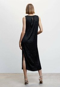 Vestido negro sin mangas con un detalle en forma de ojal en la espalda, a la altura de la rodilla, textura suave, con aberturas laterales, combinado con unos tacones plateados con tiras.
