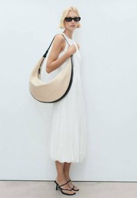 Beige tyg hobo-väska med en böjd form och svart läderdetaljer, bärs på ena axeln. Modellen bär en vit klänning och svarta solglasögon.