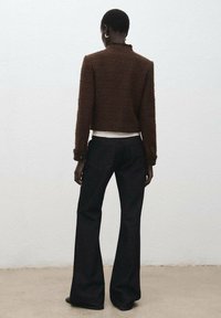 Bruine gestructureerde cropped jas met een hoge kraag, gecombineerd met zwarte wijde jeans. De jas heeft manchetten met knopen als detail.