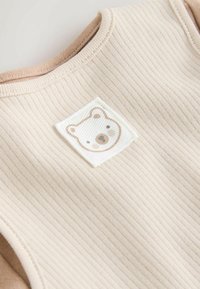 Vêtement beige côtelé avec un col marron clair. Il présente un patch en forme de tête d'ours bien visible, dans des tons doux de blanc et de marron.